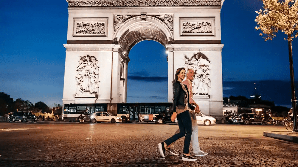 A couple walks hand-in-hand in front of the illuminated Arc de Triomphe in Paris at night, with city lights glowing around the historic monument.